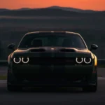 Black Dodge Challenger SRT Hellcat parked on a racetrack at sunset with glowing headlights and dramatic evening landscape wallpaper