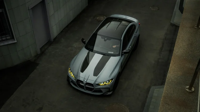 Grey BMW M3 Competition G80 viewed from above with carbon fiber accents, black roof, and glowing yellow headlights wallpaper.