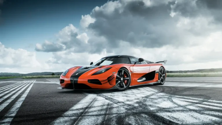 Orange Koenigsegg Agera supercar with black accents parked on an open track beneath dramatic cloudy skies wallpaper.