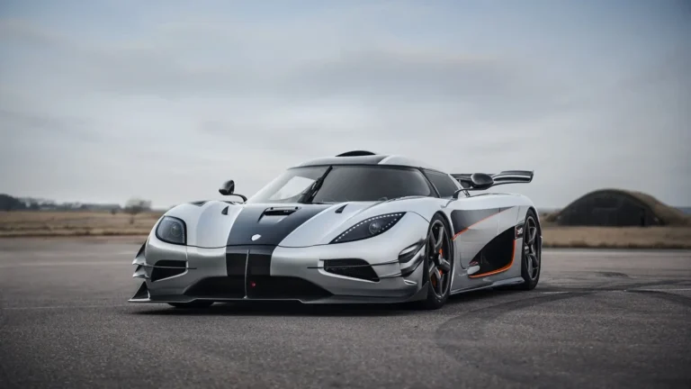 Silver Koenigsegg Agera RS hypercar with black carbon fiber racing stripes, orange accents, aerodynamic front splitter, and large rear wing parked on an empty runway under a cloudy sky wallpaper.