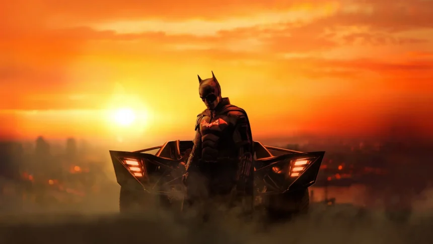The Batman 2022 Robert Pattinson in his dark Batsuit standing in front of the Batmobile with a glowing orange sunset and smoky Gotham skyline wallpaper.